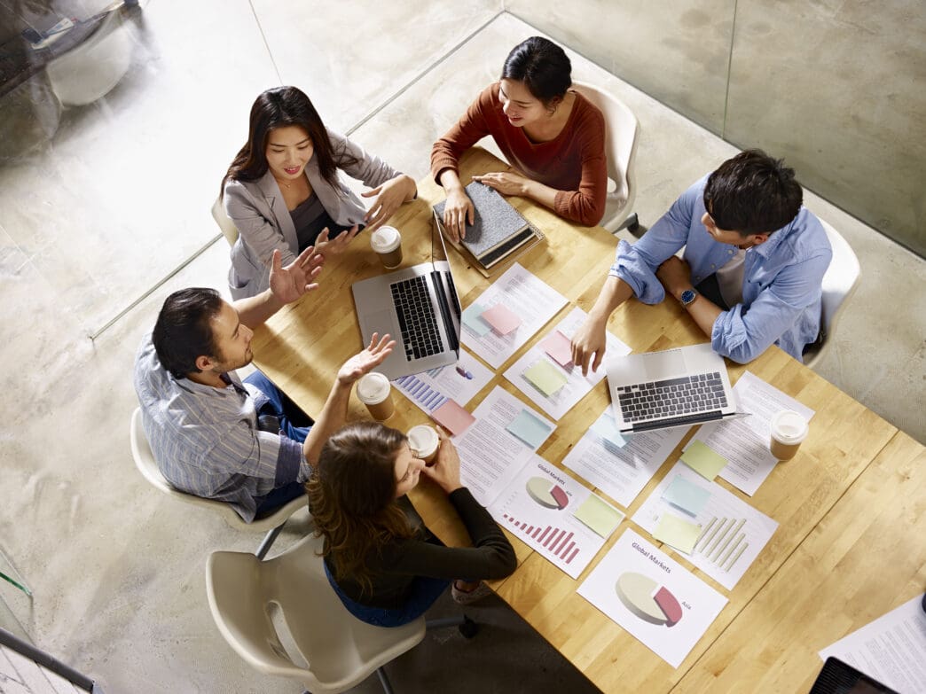 A diverse group of five Chinese professionals collaborates around a wooden table filled with documents and laptops