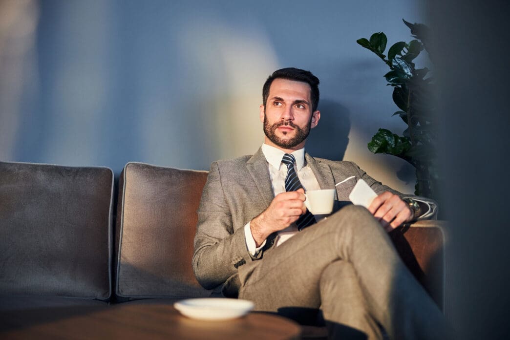 A businessman in a suit sits on a couch holding coffee, looking up thoughtfully.