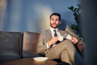 A businessman in a suit sits on a couch holding coffee, looking up thoughtfully.