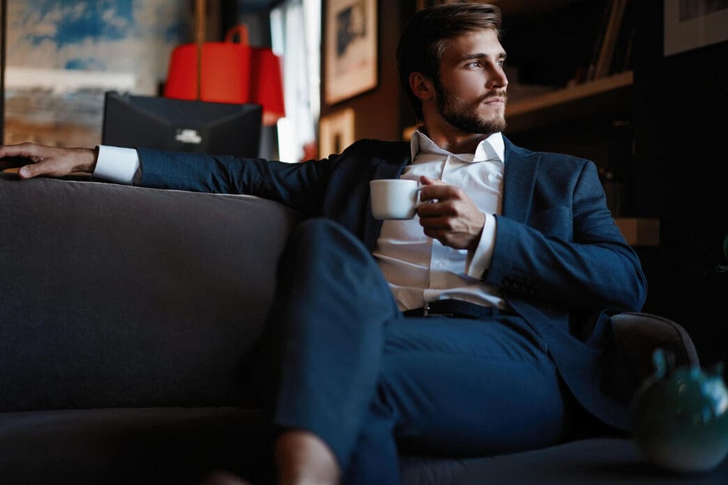 Man in a suit sits on a couch holding a cup of coffee and looking away