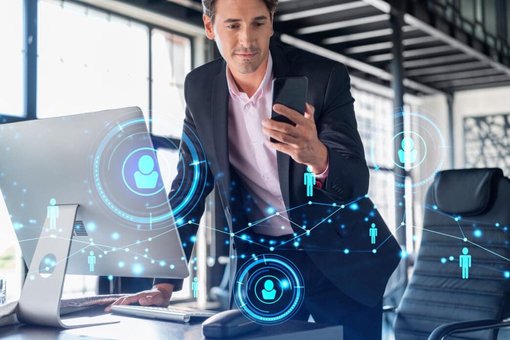 Businessman in a suit and pink shirt looking at his smartphone, with a glowing blue network overlay showing user profile icons connecting his desktop monitor and surroundings.
