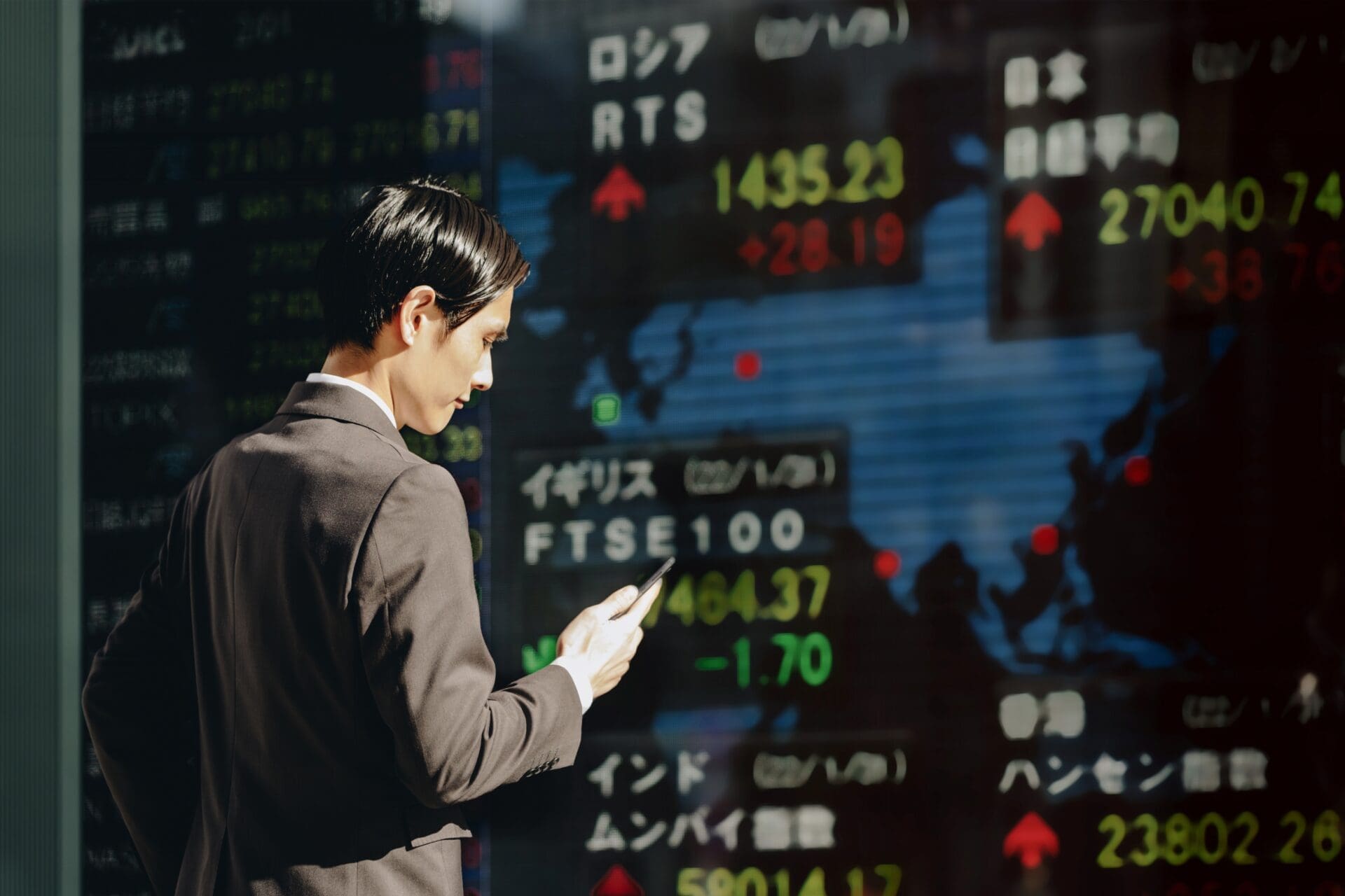 Businessman in a dark suit checks his smartphone while standing in front of a large digital screen displaying global stock market indices.