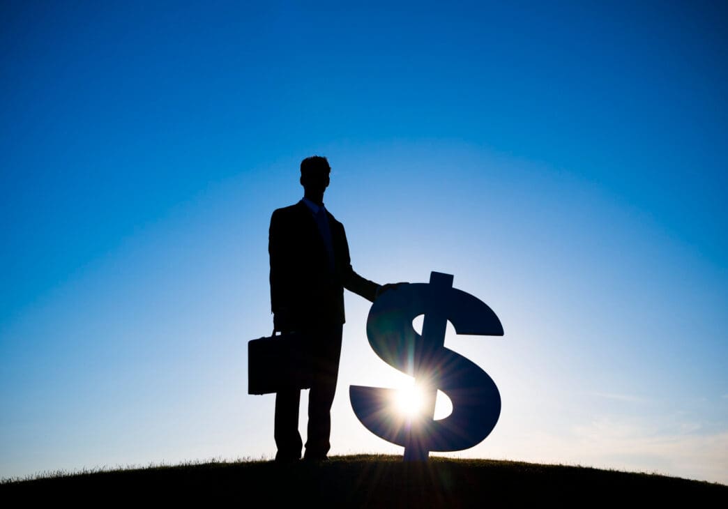 Silhouette of a businessman with a briefcase standing next to a large dollar sign against a bright blue sky