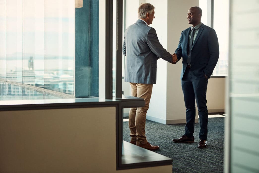 Two businessmen in suits shake hands in a modern high-rise office with large windows.