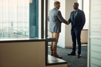 Two businessmen in suits shake hands in a modern high-rise office with large windows.