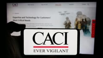 The CACI International Inc. logo on a smartphone screen, with the company's website in the background.