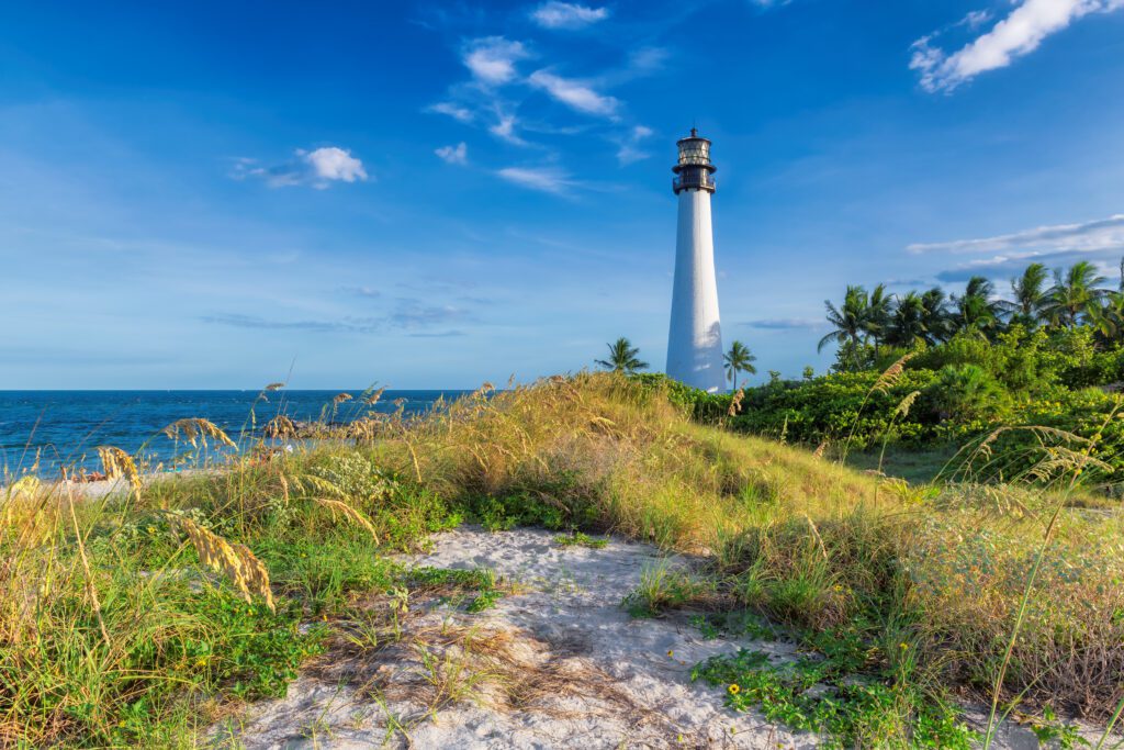 The tall, white Cape Florida Lighthouse stands on a grassy dune overlooking the Atlantic Ocean under a bright blue sky.