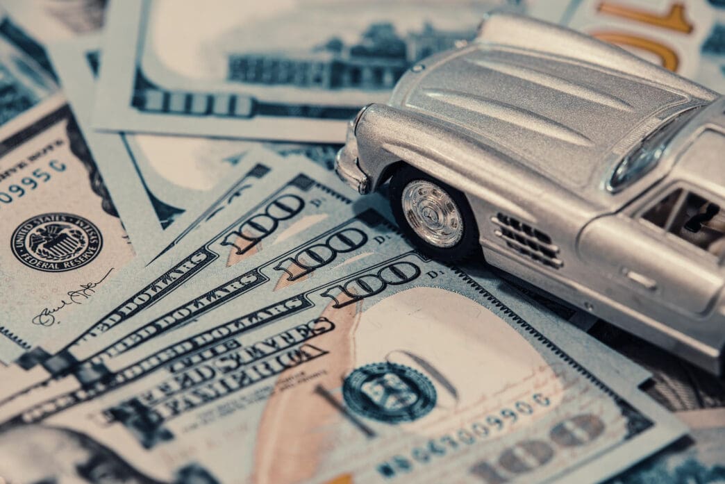 A classic silver model car sitting on top of a pile of US one-hundred-dollar bills.