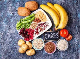 A collection of carbohydrate-rich foods, including fruits, vegetables, grains, and legumes, are arranged on a dark blue surface.