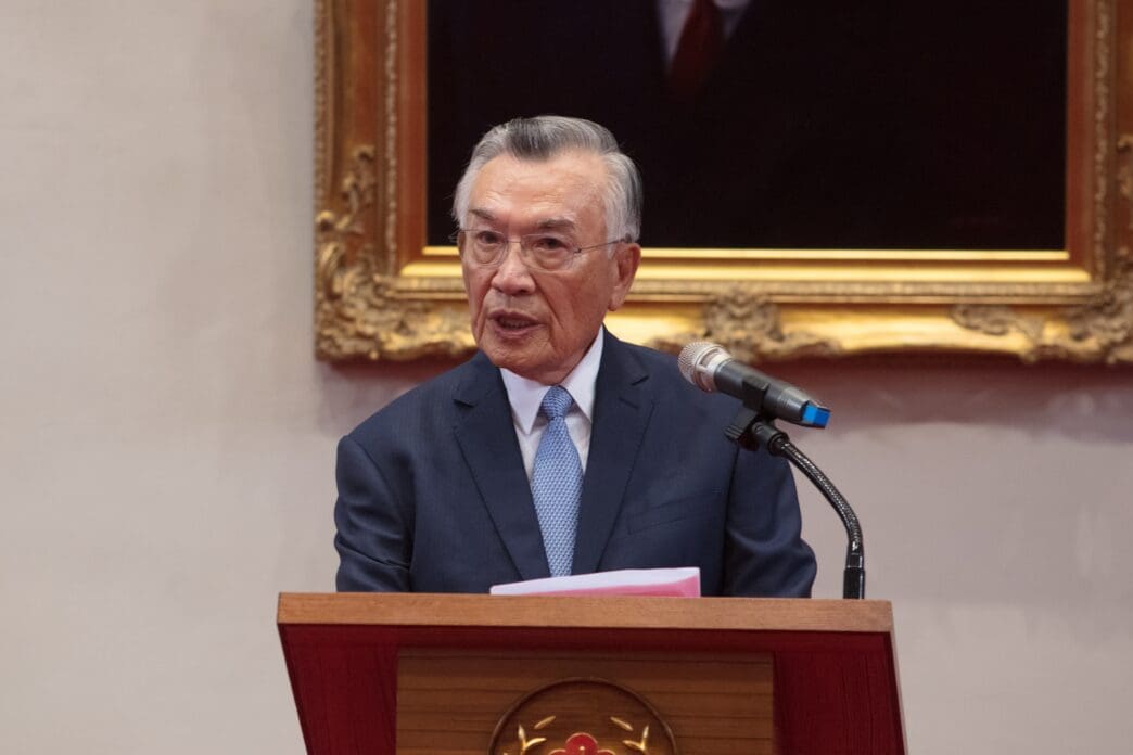Chairman Lin Hsin-i, an older man in glasses and a dark suit, speaking from a wooden podium with a microphone.