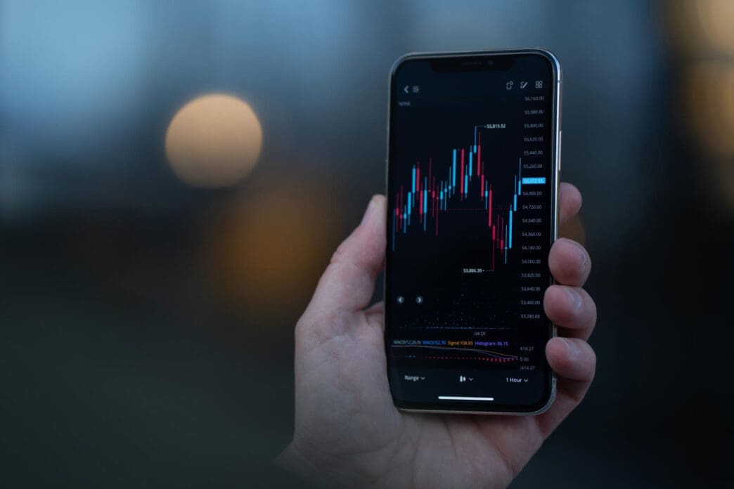 Hand holding a smartphone displaying a candlestick stock chart app against a blurred background.
