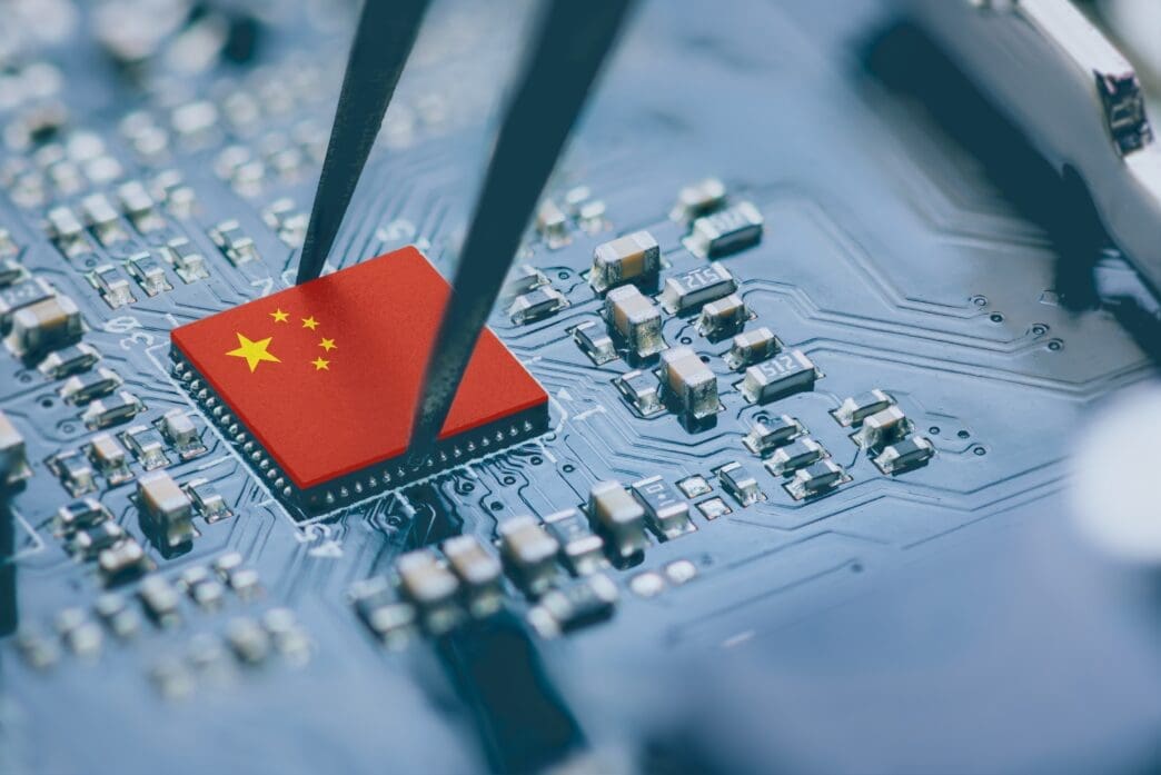 Tweezers holding a microchip bearing the Chinese flag over a dark blue circuit board filled with electronic components.