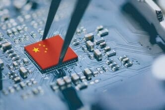 Tweezers holding a microchip bearing the Chinese flag over a dark blue circuit board filled with electronic components.