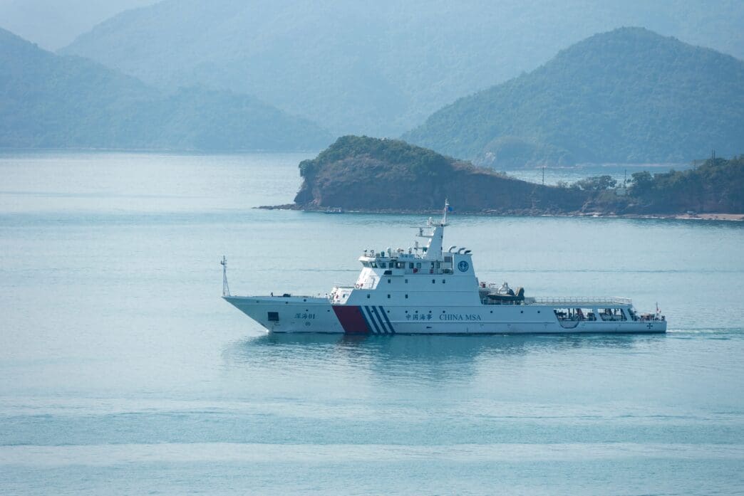 White Chinese Coast Guard ship with red and blue stripes and "CHINA MSA" text patrols calm, hazy waters with green, mountainous islands in the background.