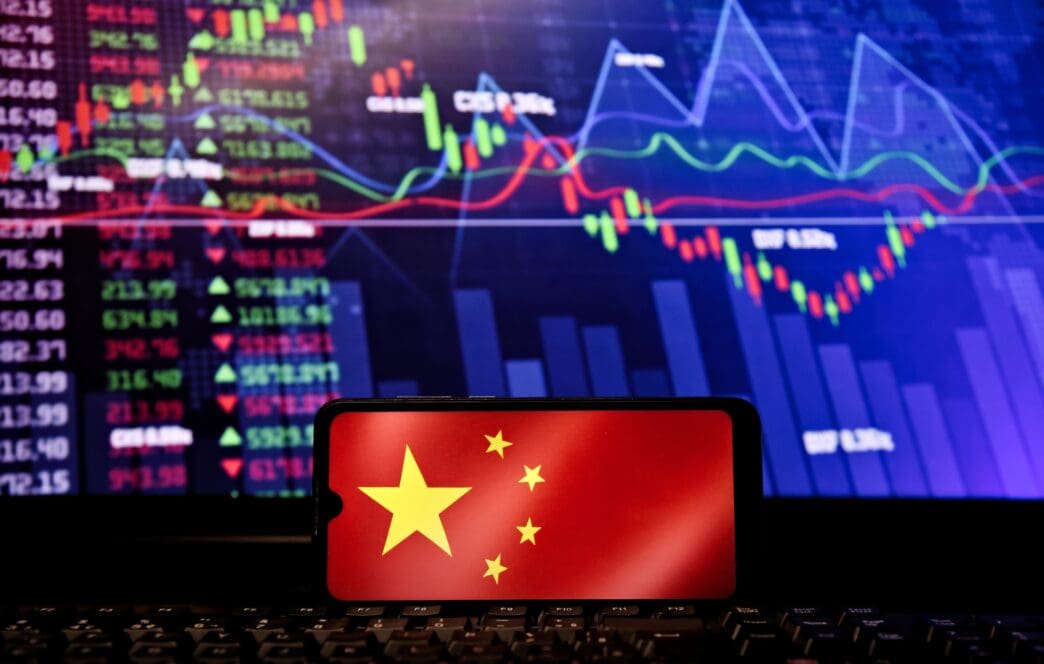 Chinese flag displayed on a phone screen in front of a monitor showing vibrant stock market charts and data.