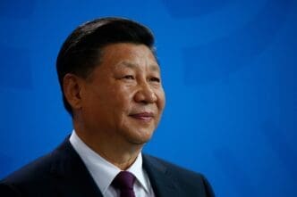 Close-up portrait of Chinese President Xi Jinping wearing a dark suit and red tie, smiling slightly against a bright blue background.