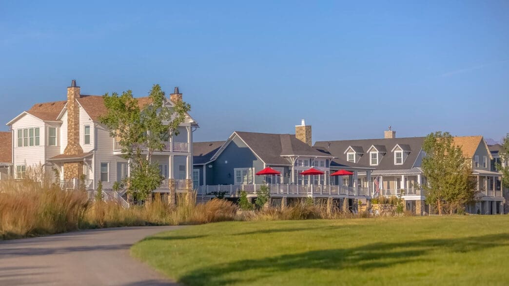 A row of large, modern coastal-style homes with porches and red umbrellas viewed from a grassy park.