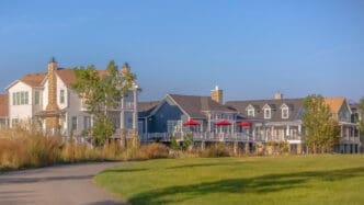 A row of large, modern coastal-style homes with porches and red umbrellas viewed from a grassy park.