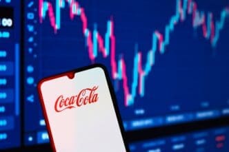A smartphone displaying the red Coca-Cola logo is held in front of a blurry, digital stock market candlestick chart.
