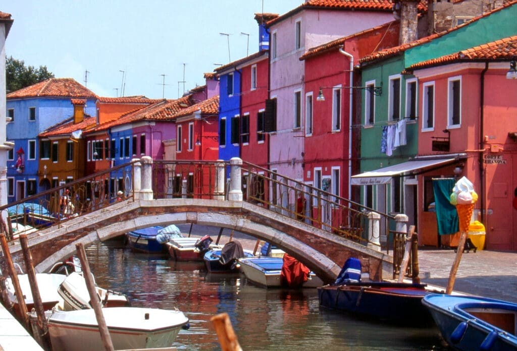 A narrow canal in Burano, Italy, with brightly painted houses on both banks and small boats moored beneath a brick bridge.