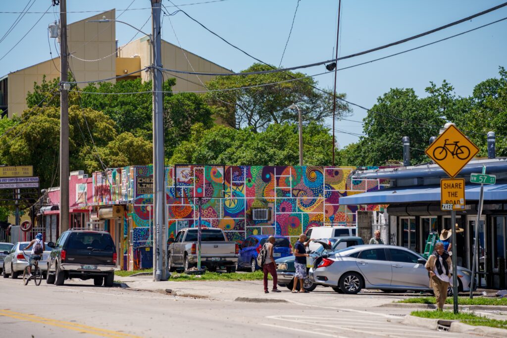 Street scene in Little Haiti, Miami, featuring a wall covered in a brightly colored mosaic mural, cars, and pedestrians.