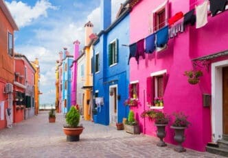 A narrow cobblestone street lined with extremely bright houses in magenta, blue, and orange under a blue sky.