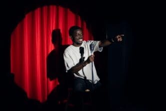 A happy African American man doing stand-up comedy on a stage with red curtains, holding a microphone and pointing to the audience.
