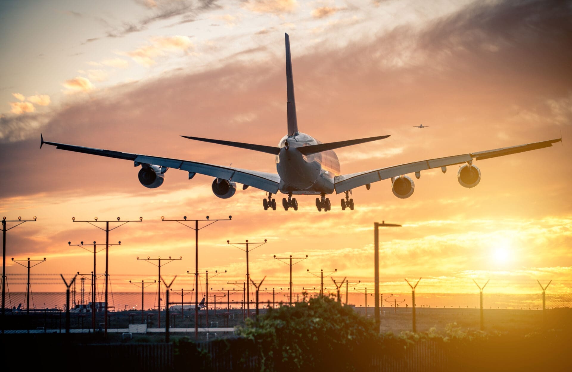 A large passenger airplane descending for a landing, viewed from the rear, silhouetted against a dramatic orange and yellow sunset