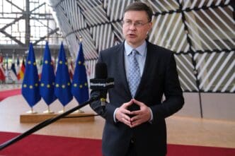 European Commissioner Valdis Dombrovskis speaks to reporters, standing in front of EU flags and a geometric wall inside the EU headquarters.