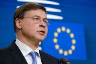 European Commissioner Valdis Dombrovskis, wearing glasses and a suit, speaks at a press conference podium against a backdrop with the EU flag's stars.