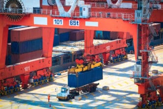 Large orange gantry crane lifting a blue shipping container off a truck trailer in a bright, sunny port terminal.
