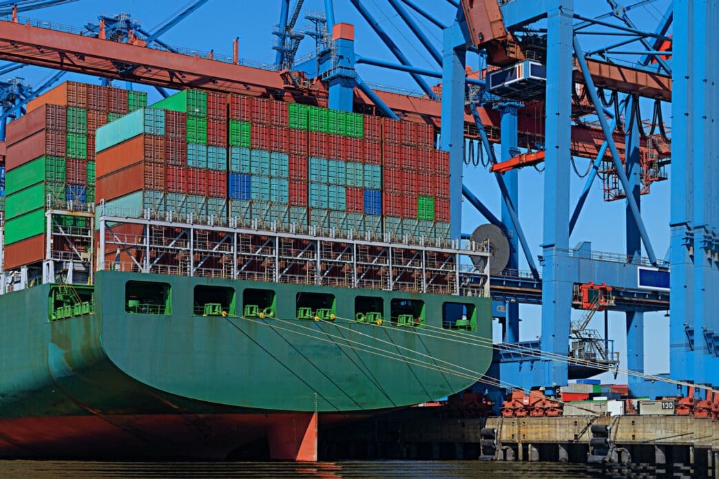 A cargo ship loaded with colorful stacked shipping containers docked beside large blue and red cranes.