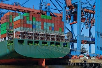 A cargo ship loaded with colorful stacked shipping containers docked beside large blue and red cranes.