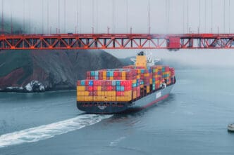 A large container ship loaded with colorful boxes sailing under the orange structure of the Golden Gate Bridge in heavy fog.