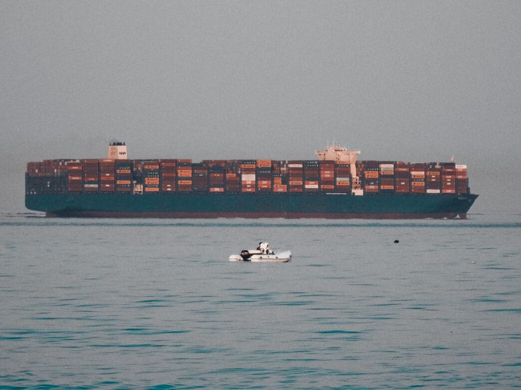 A large container ship sails on the ocean, with a tiny motorboat in front of it.