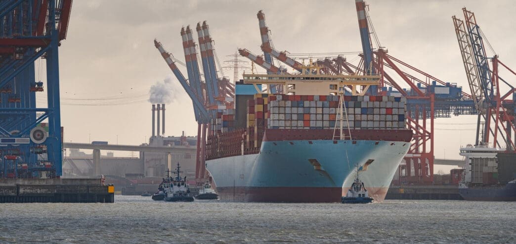 A massive container ship being guided by tugboats through a busy port with many cranes and industrial chimneys.