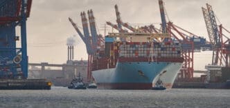 A massive container ship being guided by tugboats through a busy port with many cranes and industrial chimneys.