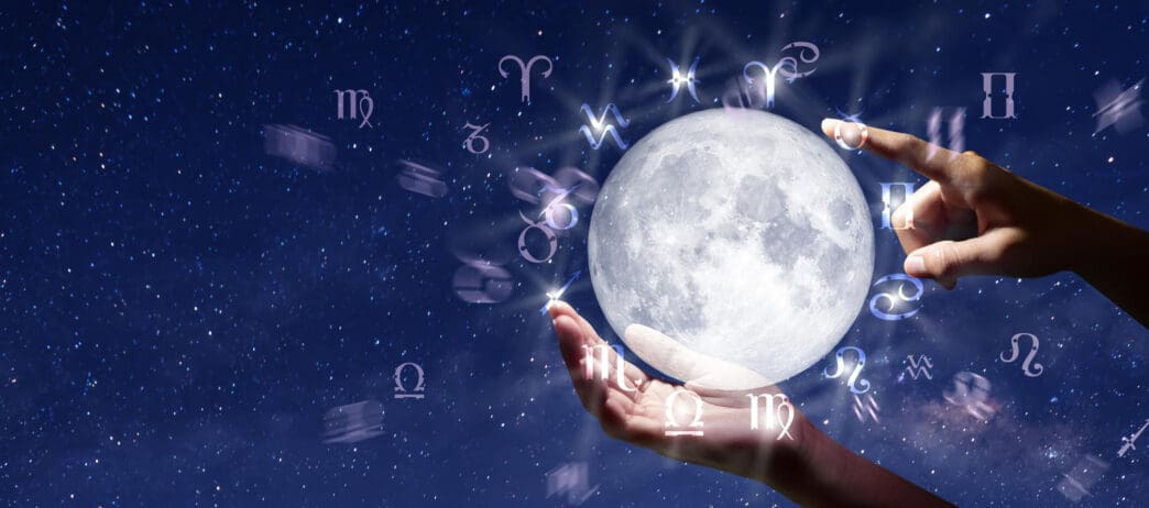Hands hold a glowing moon surrounded by zodiac symbols in a starry night sky
