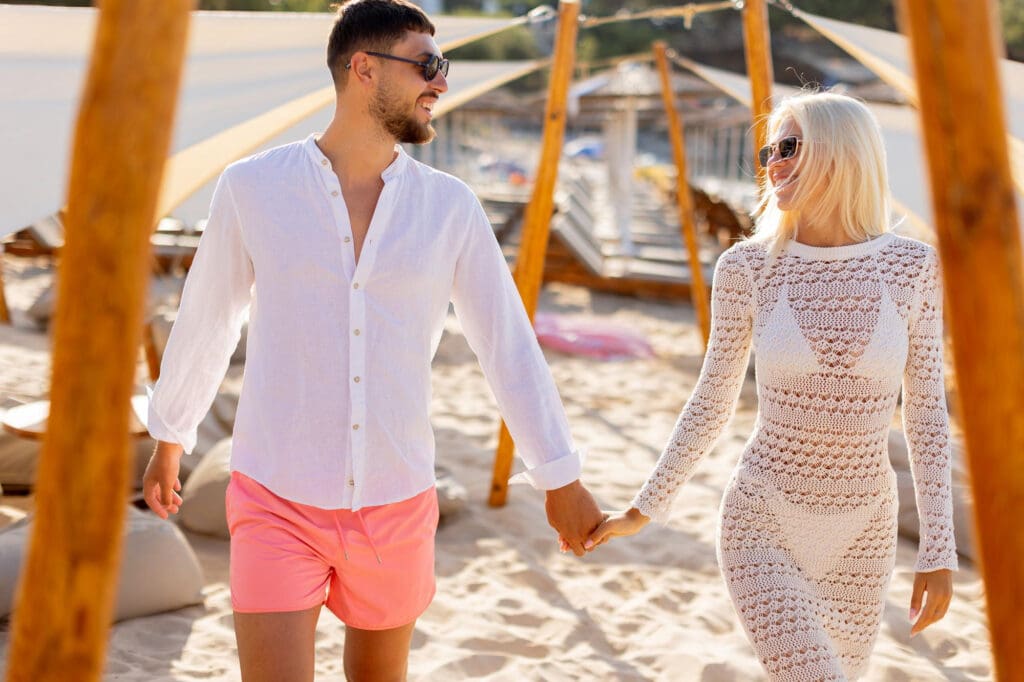 Smiling couple in stylish resort wear walks hand-in-hand on a sandy beach path lined with white cabanas.