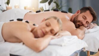 A couple relaxes side-by-side on massage tables, each with hot stones placed on their back.