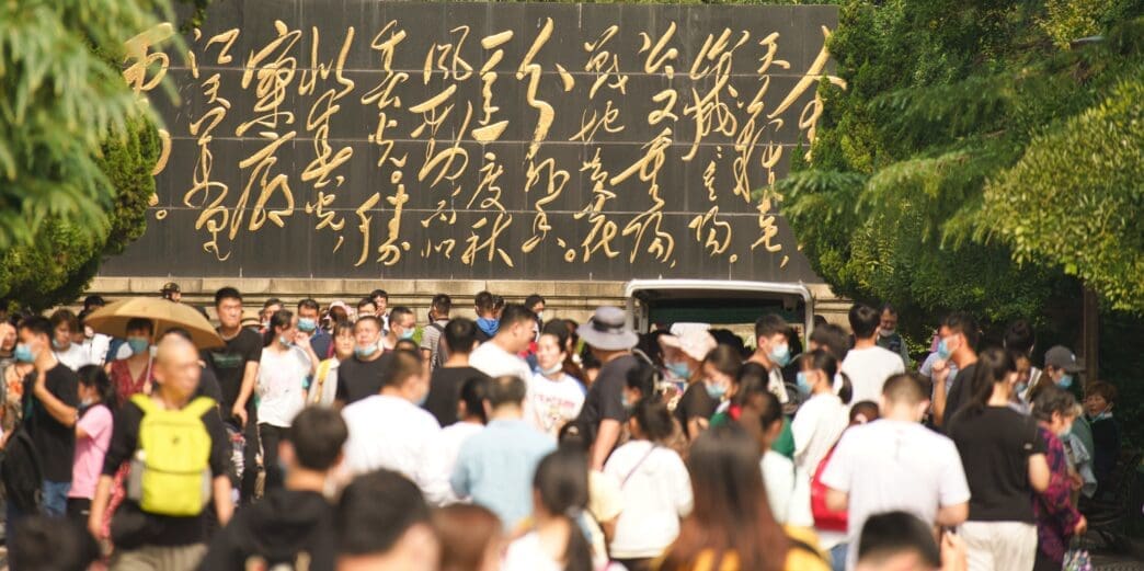 A large crowd of people walking below a black wall inscribed with prominent, gold Chinese calligraphy in a park in Jinan, China.