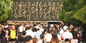 A large crowd of people walking below a black wall inscribed with prominent, gold Chinese calligraphy in a park in Jinan, China.