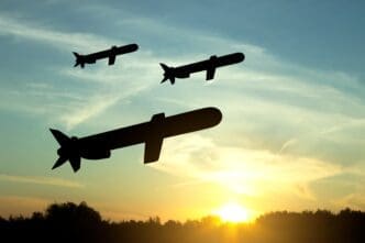 Silhouettes of three cruise missiles flying over a treeline into a pale sky during a bright orange and yellow sunset.