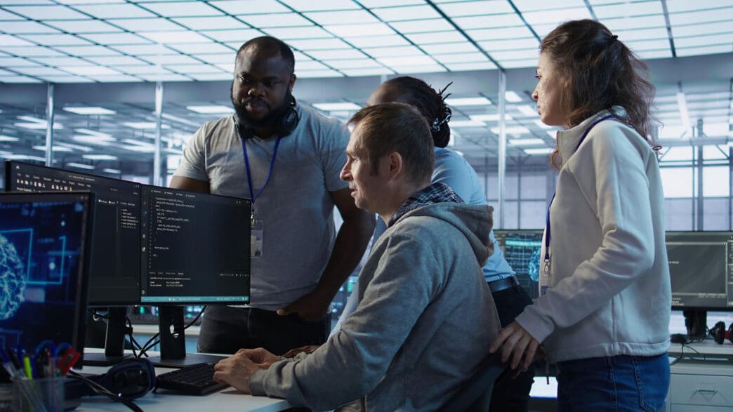 A diverse team collaborates around computer monitors displaying code in a modern office