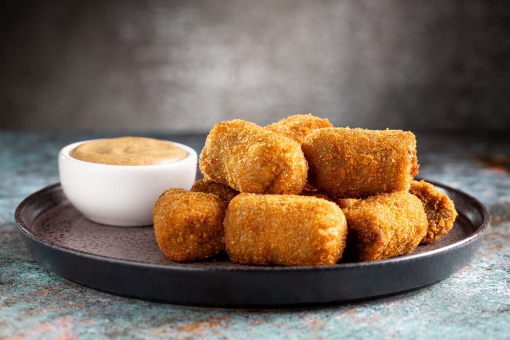 A plate of traditional fried meat croquettes piled next to a small white bowl of dipping sauce.