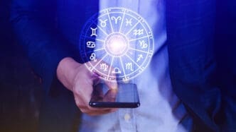 Man holding a smartphone over which a glowing, holographic zodiac wheel with all twelve astrological signs is projected.