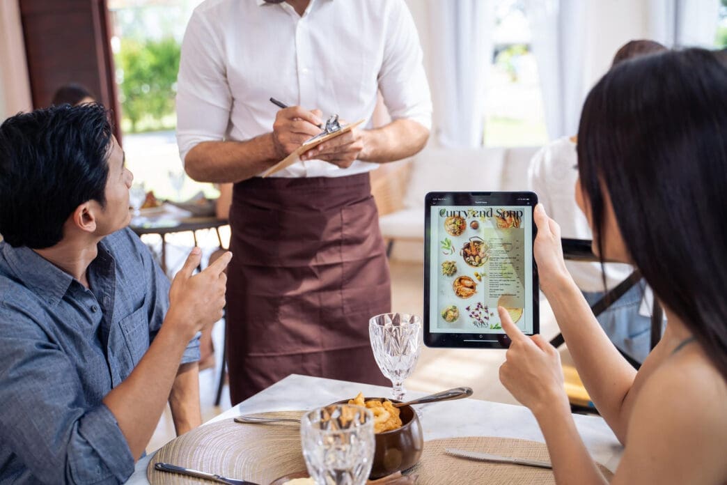 Customer points at a digital tablet displaying a restaurant menu with food pictures, while a waiter stands nearby taking notes on a notepad.