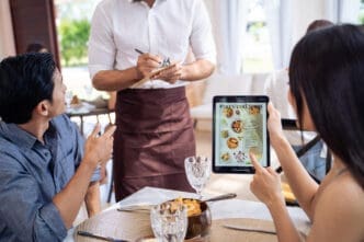 Customer points at a digital tablet displaying a restaurant menu with food pictures, while a waiter stands nearby taking notes on a notepad.