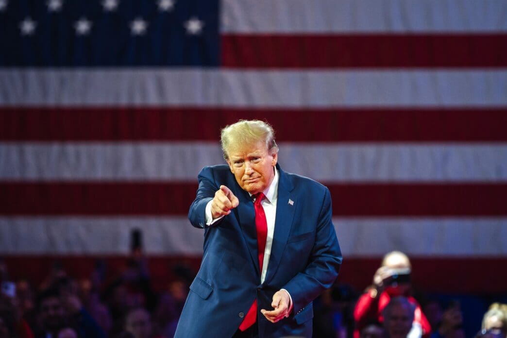 Donald Trump pointing at the camera while speaking in front of an American flag.
