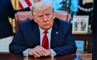 Donald Trump sits at a desk with his hands clasped, looking to the side